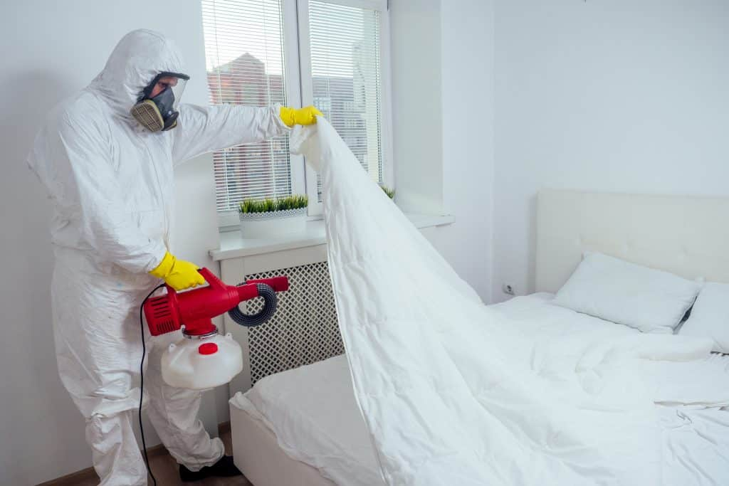 Pest control professional in full protective gear treating a bed with a fogging machine.