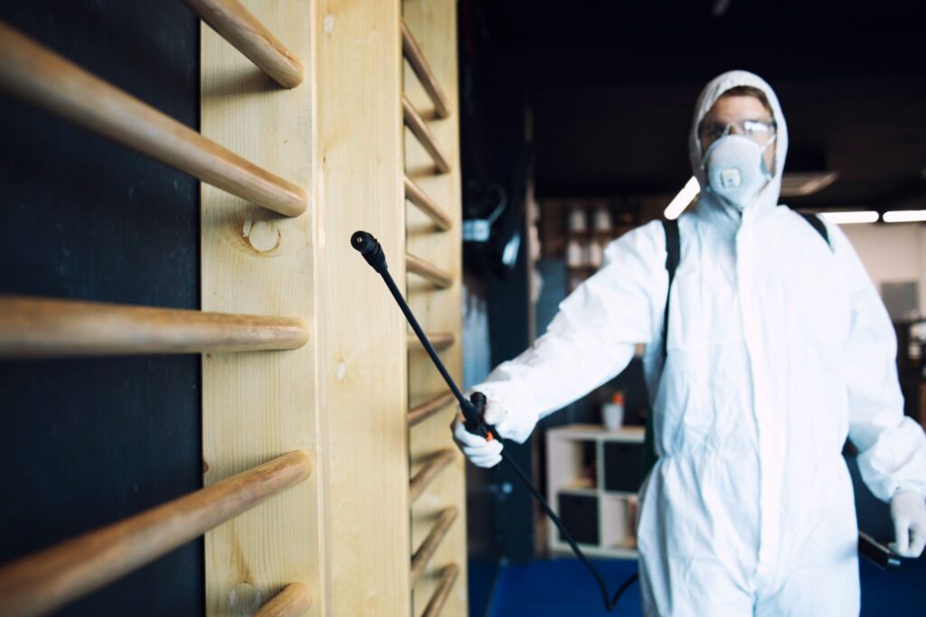 Person in white protective suit and respirator mask spraying pest control treatment.