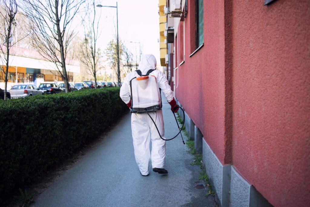 Pest control technician in a white hooded hazmat suit shown from behind walking along a sidewalk and spraying pesticide with a backpack sprayer beside a red building.