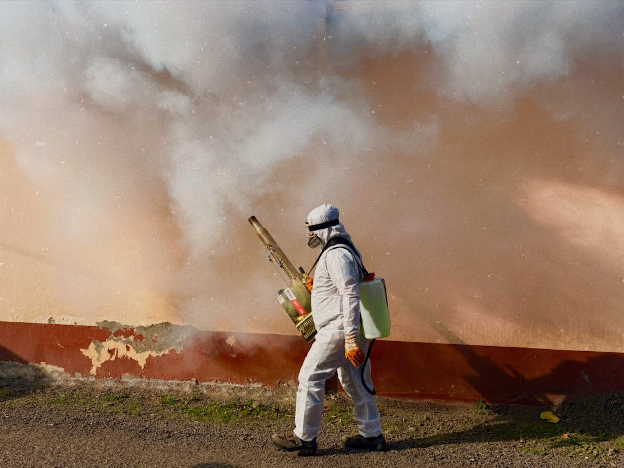 A pest control worker in protective gear uses a fogging machine, producing dense smoke outdoors.
