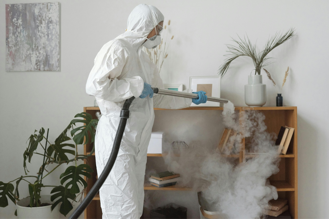 Person in protective suit and mask fumigating a living room bookshelf with steam or fog