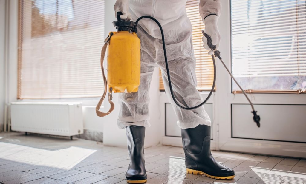 Pest control worker in a white protective suit and rubber boots holding a yellow pressure sprayer indoors.