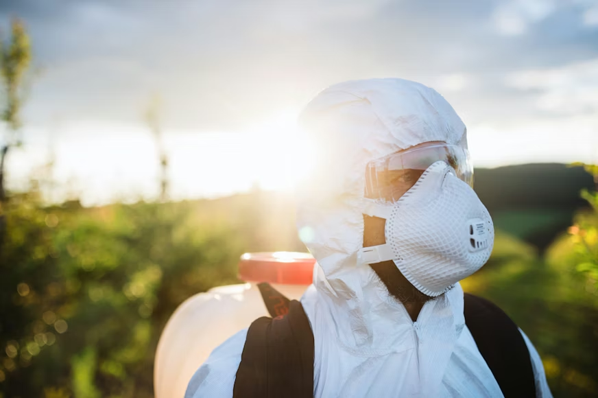Pest control technician in a white protective suit, respirator mask, and safety goggles carrying a backpack sprayer outdoors with sunlight and green foliage in the background.