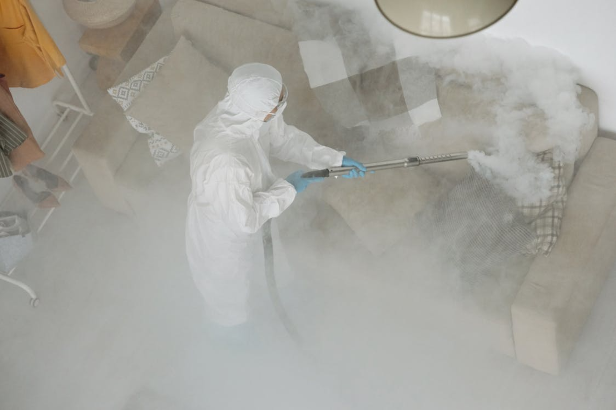 Overhead view of a pest control technician in a white hooded hazmat suit, safety goggles, face mask, and blue gloves spraying disinfectant fog onto a beige sofa.