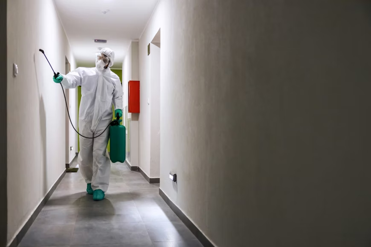 Pest control technician in a white hooded hazmat suit, respirator mask, and green gloves and boot covers walking down a hallway holding a green pressure sprayer with the nozzle raised