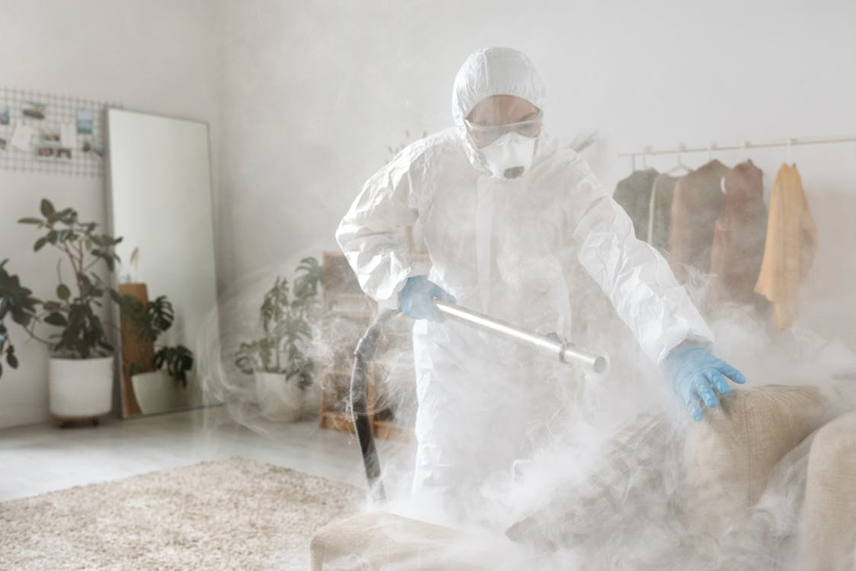 Pest control technician in a white hooded hazmat suit, safety goggles, face mask, and blue gloves spraying disinfectant fog onto a sofa, with dense white mist filling the room.