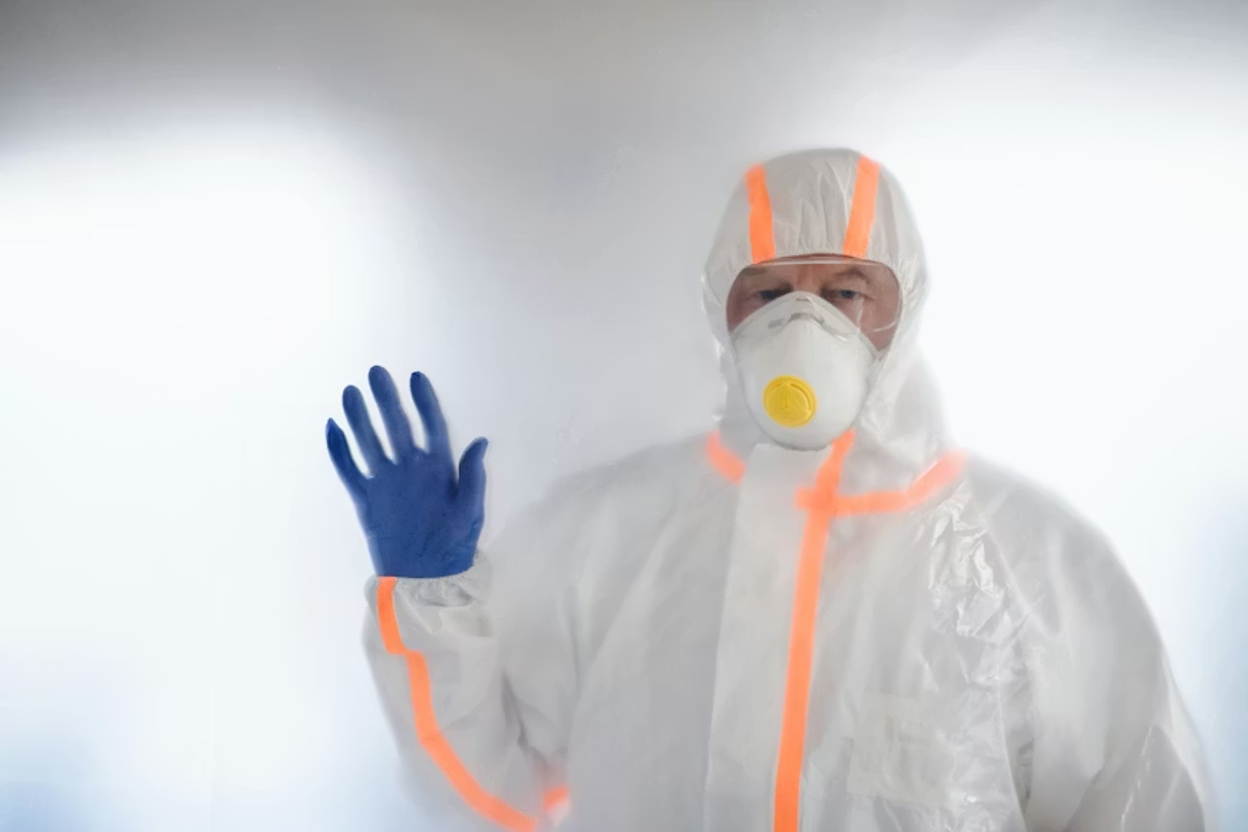 Pest control technician in a white hooded hazmat suit with orange trim, a yellow-valved respirator mask, and blue gloves raising one hand