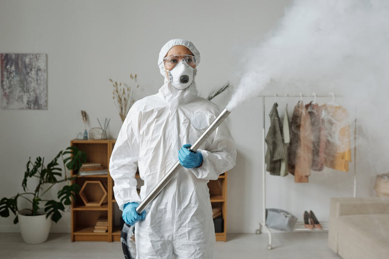 Pest control technician in a white hooded hazmat suit, safety goggles, respirator mask, and blue gloves standing and holding a metal fogging wand emitting white disinfectant mist.
