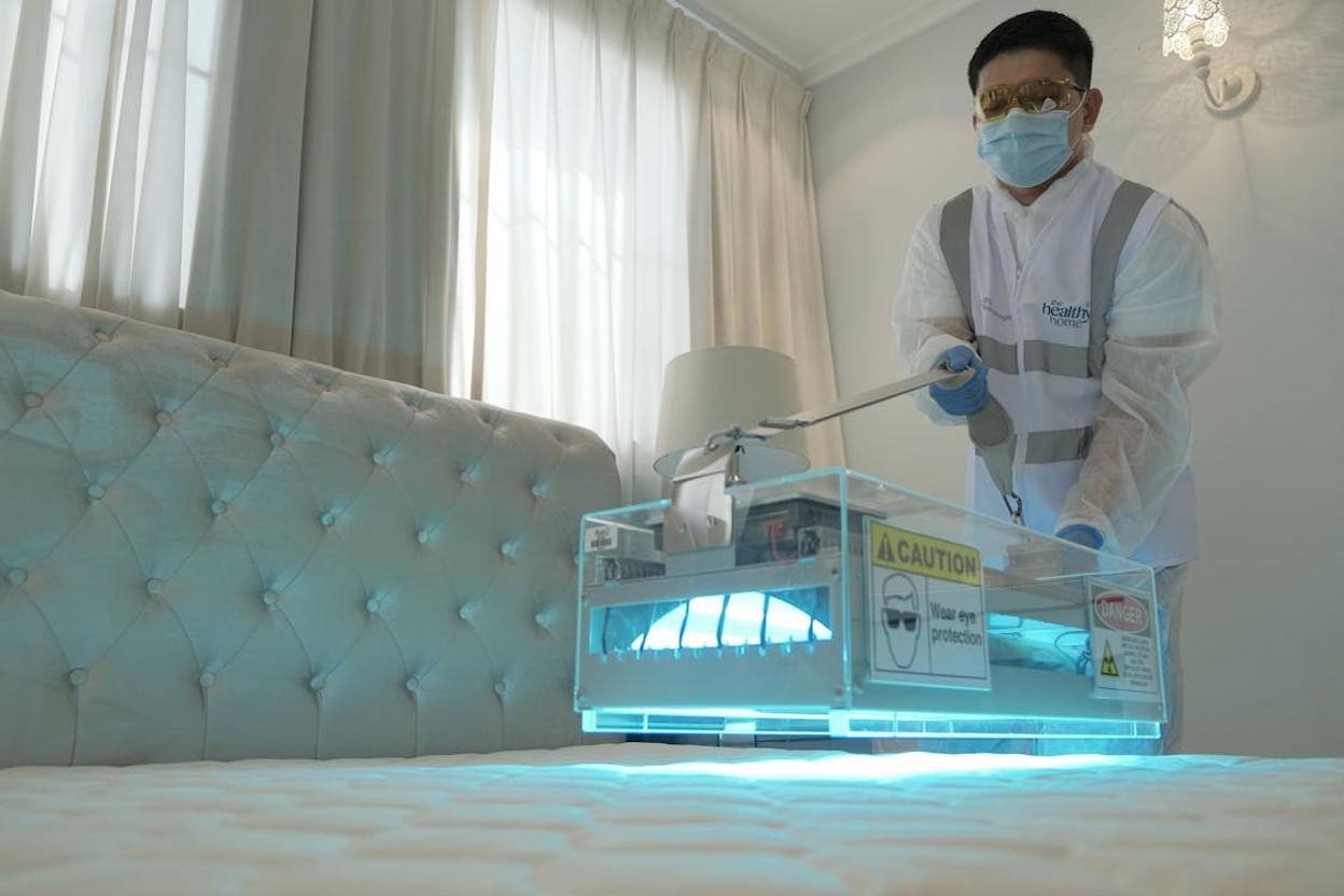 Pest control technician in safety goggles, a face mask, blue gloves, and a Healthy Home-branded high-visibility vest using a UV light sanitization device on a mattress