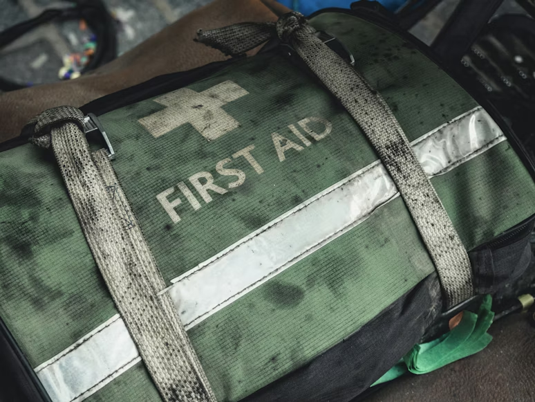 Close-up of a worn green First Aid bag with a white cross emblem, reflective stripes, and canvas carrying straps, with other equipment partially visible in the softly blurred background. 