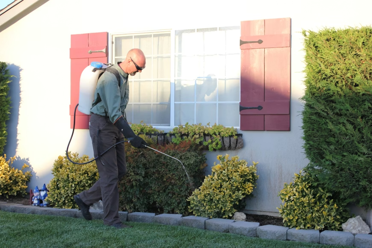 pest control technician in sunglasses, a green long-sleeved shirt, and dark overalls spraying pesticide along the foundation of a house with a backpack sprayer.