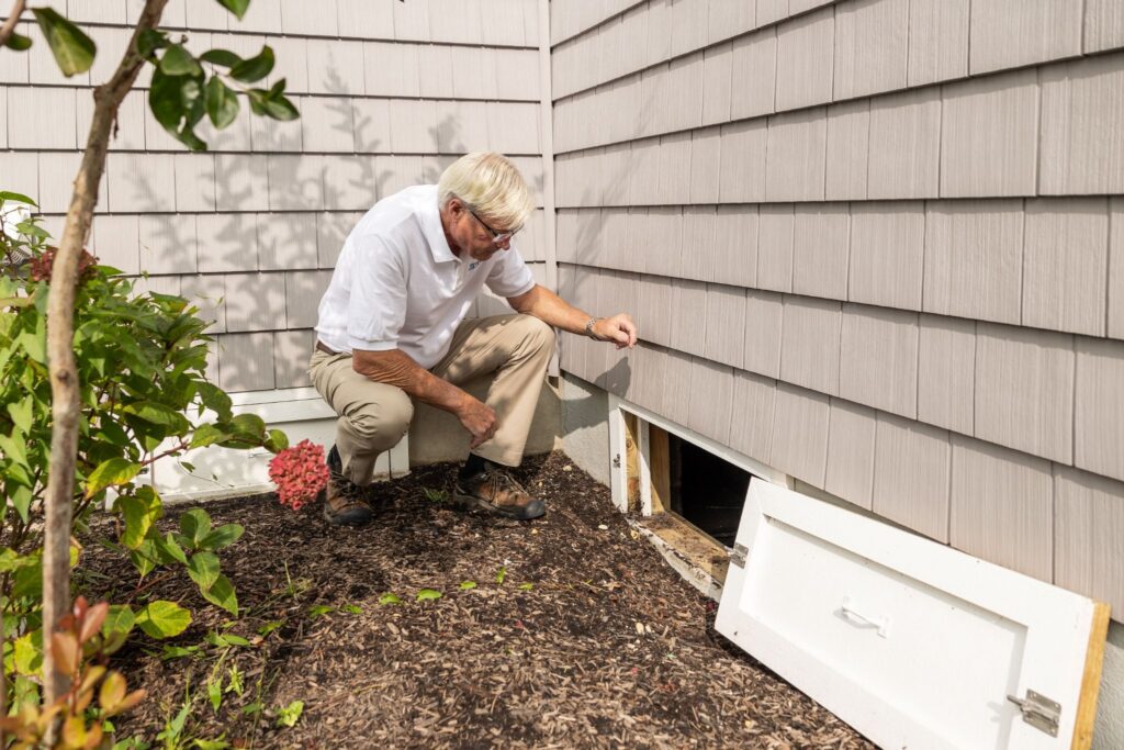 Pest control technician crouching to inspect a home's siding and open crawl space.