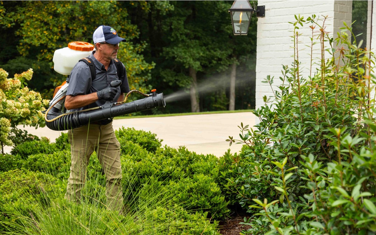 Uniformed pest control technician applying mosquito barrier treatment to residential garden shrubs with a backpack fogger