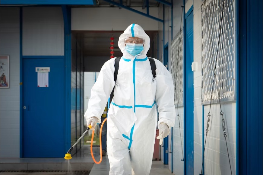 Pest control technician in a white hooded hazmat suit, safety goggles, face mask, and white gloves holding a sprayer wan