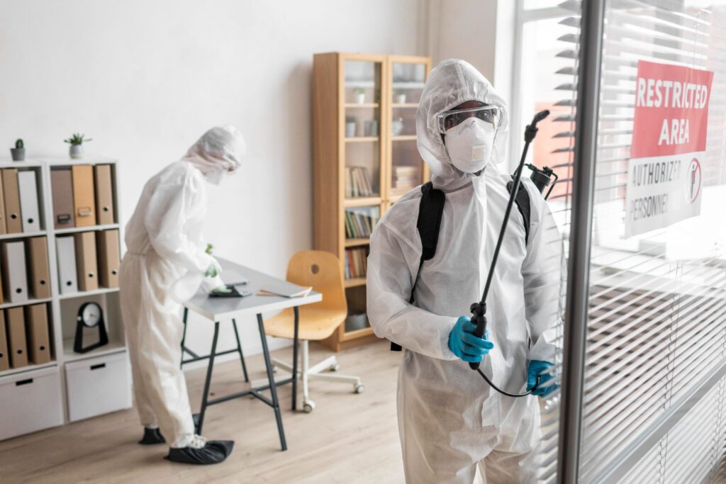 Two workers in white protective suits disinfect an office, one spraying near a window marked "Restricted Area”.