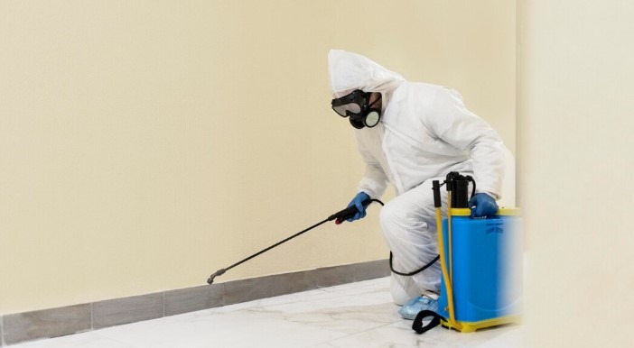 Pest control technician in a white protective suit and respirator, spraying insecticide along a baseboard.