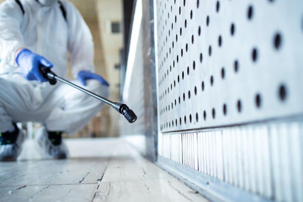 Pest control professional in protective gear applying targeted spray treatment along a baseboard and ventilation panel.