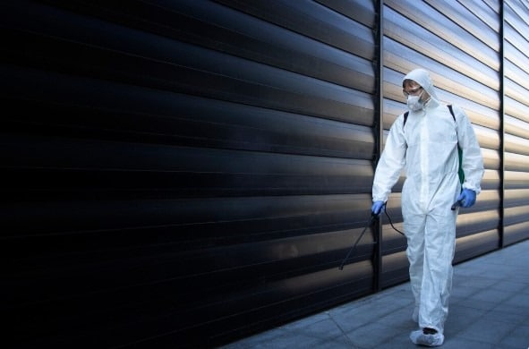 A pest control worker in a full protective suit sprays treatment along an exterior wall.