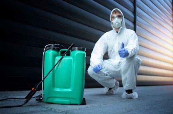 A pest control technician in a white protective suit, mask, and blue gloves crouching next to a green sprayer tank and giving a thumbs-up.