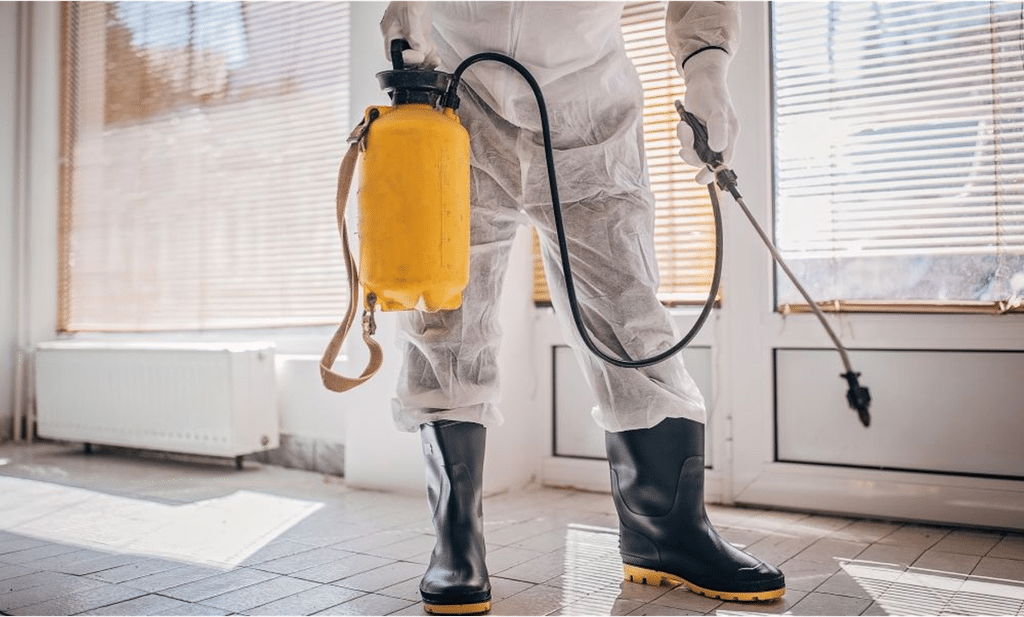 Pest control technician in protective suit and rubber boots spraying treatment indoors with a yellow pump sprayer.